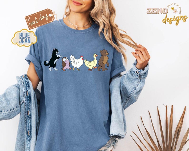 May include: Blue jean t-shirt featuring a line of cartoon animals: a black and white cat, an owl, a white chicken, a yellow duck, and a brown bear. The shirt has the text "next day" and "BLUE JEAN" on it. The shirt is from "ZEND designs".
