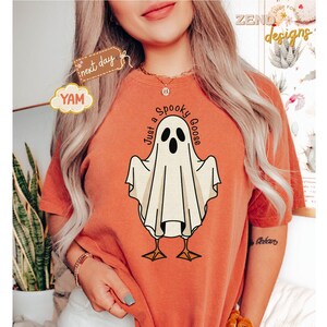 May include: Orange t-shirt featuring a cartoon ghost goose with the text "Just a Spooky Goose". The ghost is white with black eyes and a beak, standing on yellow feet. The shirt has a relaxed fit.