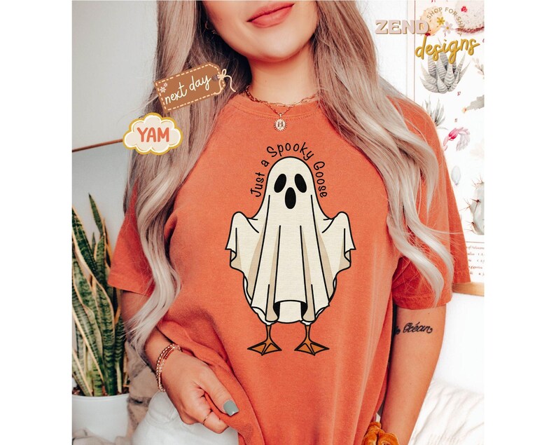 May include: Orange t-shirt featuring a cartoon ghost goose with the text "Just a Spooky Goose". The ghost is white with black eyes and a beak, standing on yellow feet. The shirt has a relaxed fit.