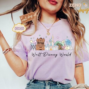 May include: A light purple t-shirt featuring a colorful illustration of Walt Disney World landmarks, including Cinderella's castle and Spaceship Earth. The words "Walt Disney World" are written in a script font below the illustration.