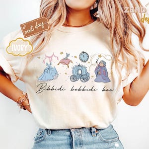 May include: Ivory-colored t-shirt featuring a whimsical illustration of a fairytale theme. The design includes a blue gown, a carriage, a clock, and a fairy godmother, with the phrase "Bibbidi Bobbidi Boo" in script. The shirt has a relaxed fit.