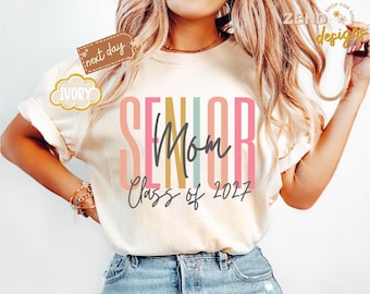 Op de afbeelding: Een ivoorkleurig t-shirt met de tekst SENIOR in grote, verticale, veelkleurige blokletters in tinten perzik, geel, blauwgroen en roze. Daaronder staan het woord Mom en de tekst Class of 2027 gedrukt in een donkergrijs cursief handschrift.