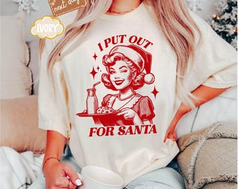 May include: A white t-shirt with red text that reads "I put out for Santa". The text is in a retro font and is accompanied by a vintage illustration of a woman in a Santa hat holding a tray with a glass of milk and a cookie.
