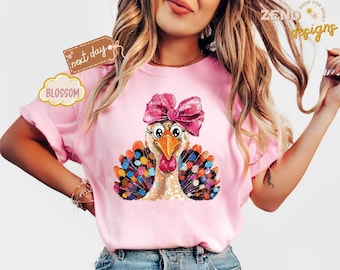 Könnte beinhalten: Rosa T-Shirt mit einem Cartoon-Truthahn-Design. Der Truthahn hat eine rosa Schleife, eine Brille und bunte Federn. Auf dem Shirt sind auch das Wort "Blossom" und der Text "next day" zu sehen.