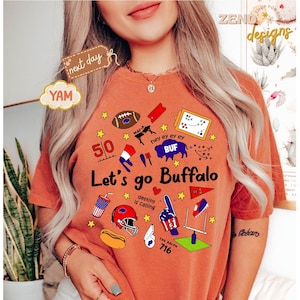 May include: Burnt orange t-shirt with the text "Let's go Buffalo" and various sports-themed graphics, including a football, helmet, and goalpost. The shirt also features the text "destiny is calling" and "The Ralph 716".