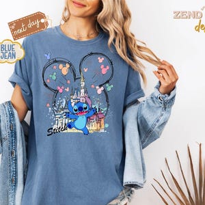 May include: Blue jean-colored t-shirt featuring a graphic of Stitch from Lilo & Stitch, with a castle and Mickey Mouse ears design. The word "Stitch" is written in black script. The shirt has rolled-up sleeves.