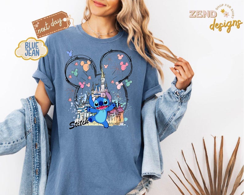 May include: Blue jean-colored t-shirt featuring a graphic of Stitch from Lilo & Stitch, with a castle and Mickey Mouse ears design. The word "Stitch" is written in black script. The shirt has rolled-up sleeves.