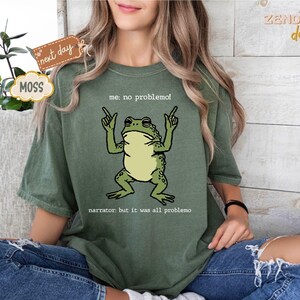 Puede incluir: Camiseta verde musgo con un dibujo de una rana de dibujos animados y el texto "me: no problemo!" y "narrator: but it was all problemo". La rana es verde y beige. La camiseta es de corte holgado.
