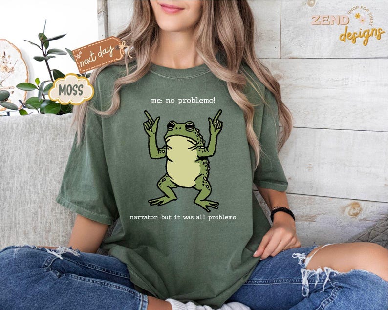 Puede incluir: Camiseta verde musgo con un dibujo de una rana de dibujos animados y el texto "me: no problemo!" y "narrator: but it was all problemo". La rana es verde y beige. La camiseta es de corte holgado.