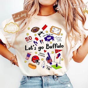 May include: Ivory-colored t-shirt with a colorful graphic design featuring football-related elements and the text "Let's go Buffalo." The design includes a football, helmet, and other sports-themed illustrations. The shirt has short sleeves.