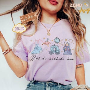 May include: A light purple t-shirt featuring a Cinderella-themed design with a dress, carriage, clock, and fairy godmother illustration. The phrase "Bibbidi Bobbidi Boo" is written in cursive. The shirt has the word "Orchid" on a tag.
