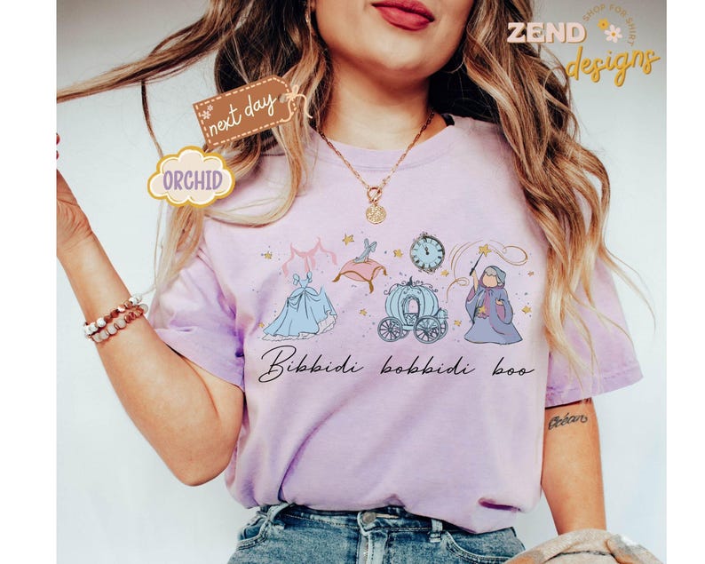 May include: A light purple t-shirt featuring a Cinderella-themed design with a dress, carriage, clock, and fairy godmother illustration. The phrase "Bibbidi Bobbidi Boo" is written in cursive. The shirt has the word "Orchid" on a tag.