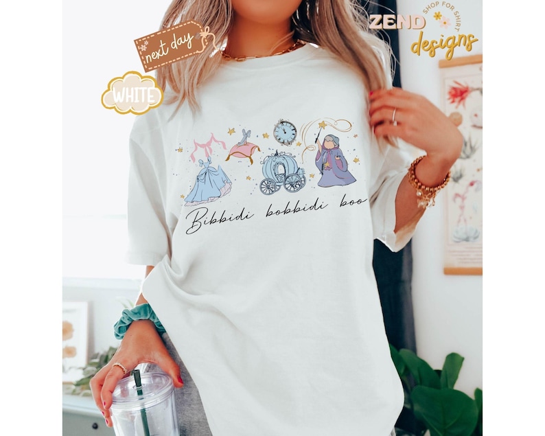 May include: White t-shirt with a whimsical illustration featuring a princess, carriage, clock, and fairy godmother. The phrase "Bibbidi Bobbidi Boo" is written in script below the design. The shirt has a relaxed fit.