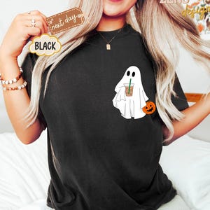 Pu&ograve; includere: Maglietta nera con un fantasma bianco che tiene una tazza di caff&egrave; e una piccola zucca arancione. Il testo "next day" &egrave; visibile nell'immagine.