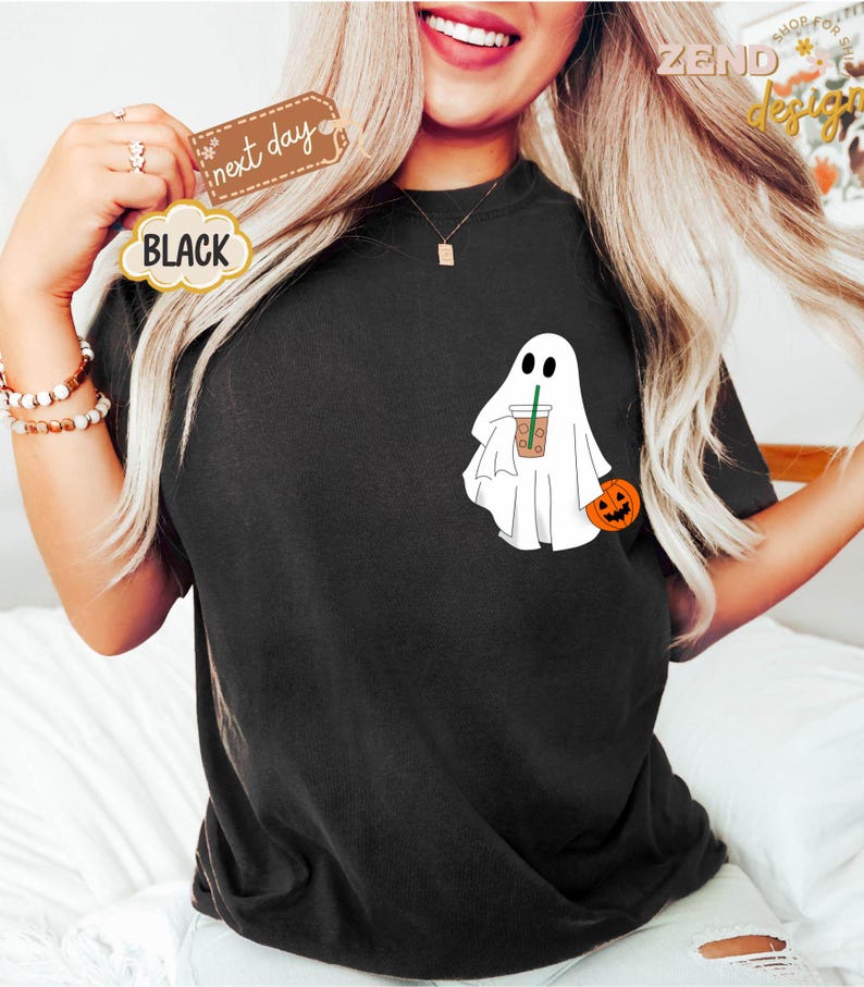Pu&ograve; includere: Maglietta nera con un fantasma bianco che tiene una tazza di caff&egrave; e una piccola zucca arancione. Il testo "next day" &egrave; visibile nell'immagine.