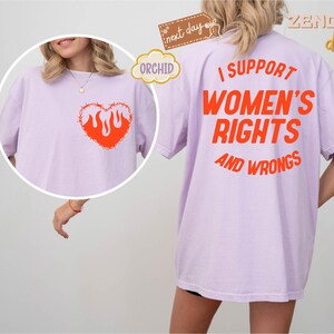 May include: A lavender t-shirt with the text "I support women's rights and wrongs" printed in orange on the back. The front of the shirt features a barbed wire heart with flames inside, also in orange.