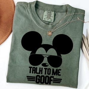 Puede incluir: Una camiseta de cuello redondo color verde oliva con un gr&aacute;fico negro de la silueta de un rat&oacute;n con gafas de aviador. El texto debajo del gr&aacute;fico dice TALK TO ME GOOF con l&iacute;neas horizontales. La camiseta tiene una etiqueta Comfort Colors.