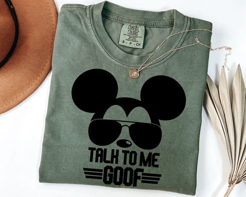 Puede incluir: Una camiseta de cuello redondo color verde oliva con un gr&aacute;fico negro de la silueta de un rat&oacute;n con gafas de aviador. El texto debajo del gr&aacute;fico dice TALK TO ME GOOF con l&iacute;neas horizontales. La camiseta tiene una etiqueta Comfort Colors.