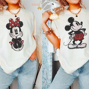 May include: Two women wearing cream-colored t-shirts with cartoon images of Mickey Mouse and Minnie Mouse. The shirts have a distressed look and the images are in black and white with red accents.