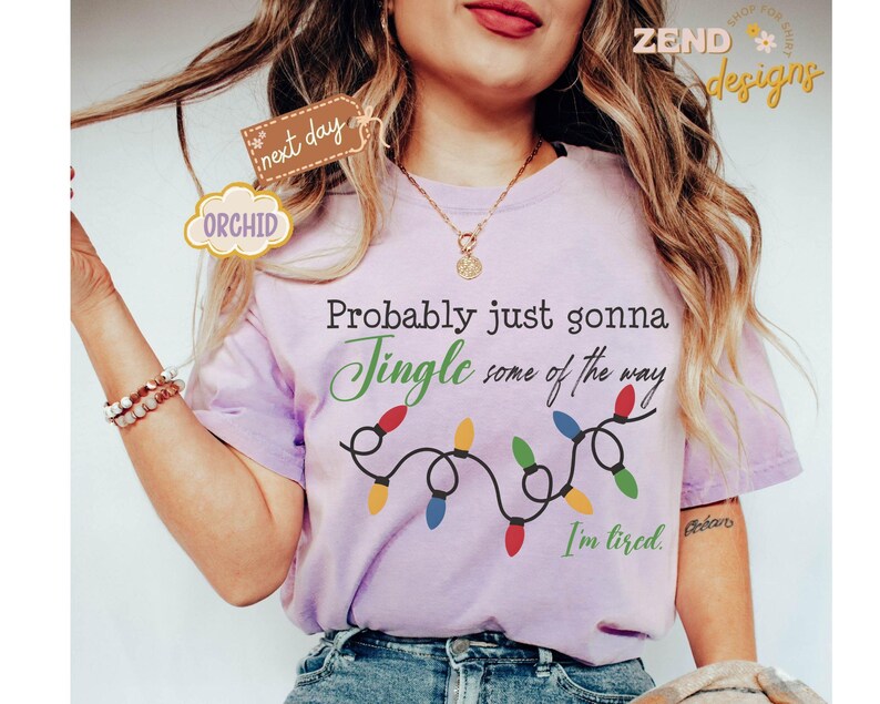 Pode incluir: Uma t-shirt lil&aacute;s clara com o texto "Probably just gonna Jingle some of the way" em preto e verde. Uma sequ&ecirc;ncia de luzes de Natal coloridas est&aacute; abaixo do texto. As palavras "I'm tired" est&atilde;o escritas em preto no canto inferior direito.