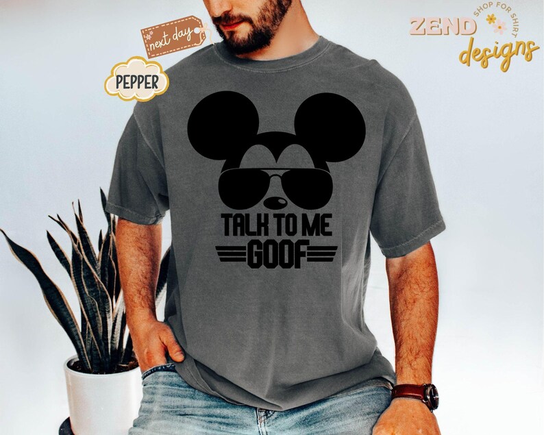 Puede incluir: Una camiseta de manga corta gris carb&oacute;n con un gr&aacute;fico negro de la silueta de un rat&oacute;n con gafas de sol. Debajo del gr&aacute;fico, el texto dice TALK TO ME GOOF en letras may&uacute;sculas negras en negrita con l&iacute;neas horizontales a ambos lados de GOOF.