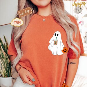 Pu&ograve; includere: Una maglietta color arancio bruciato con un fantasma bianco che tiene un bicchiere di caff&egrave; freddo marrone e una piccola zucca arancione. Il testo "next day" &egrave; su un'etichetta marrone e il testo "YAM" &egrave; su una nuvola gialla.