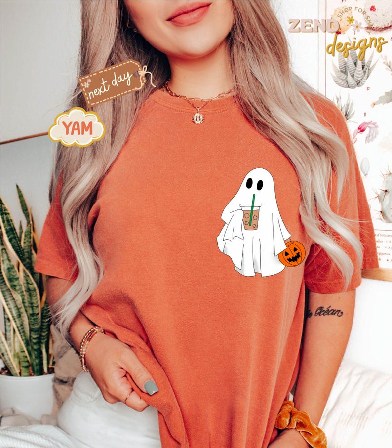 Pu&ograve; includere: Una maglietta color arancio bruciato con un fantasma bianco che tiene un bicchiere di caff&egrave; freddo marrone e una piccola zucca arancione. Il testo "next day" &egrave; su un'etichetta marrone e il testo "YAM" &egrave; su una nuvola gialla.