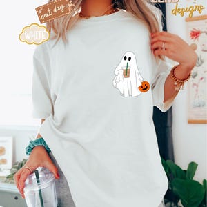 Pu&ograve; includere: Maglietta bianca con un fantasma dei cartoni animati che tiene una tazza di caff&egrave; e una piccola zucca. Il testo "ZEND designs" &egrave; visibile sullo sfondo.