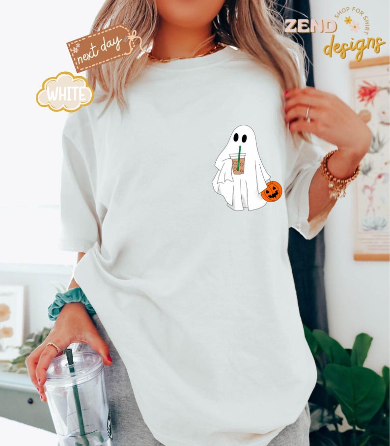 Pu&ograve; includere: Maglietta bianca con un fantasma dei cartoni animati che tiene una tazza di caff&egrave; e una piccola zucca. Il testo "ZEND designs" &egrave; visibile sullo sfondo.