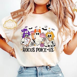 May include: Ivory-colored t-shirt featuring a Halloween-themed graphic. The design includes three cartoon ghosts dressed as nurses, holding medical items, with the text "Hocus Poke-Us" below. The shirt also has a "next day" tag.