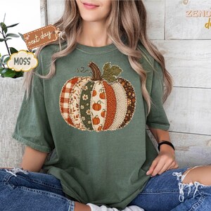 以下が含まれることがあります： モスグリーンの半袖Tシャツ。中央には、チェック柄、花柄、ヘリンボーン柄など、オレンジ、ブラウン、クリーム色の様々な縦縞の布地風パターンで構成された大きなカボチャのグラフィックがプリントされています。