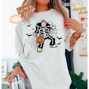 K&ouml;nnte beinhalten: Wei&szlig;es T-Shirt mit einem Halloween-Design. Die Grafik zeigt eine Figur in einem Skelettkost&uuml;m, die einen K&uuml;rbis h&auml;lt. Schwarze Flederm&auml;use und ein Spinnennetz vervollst&auml;ndigen das gruselige Design. Auf dem Shirt steht das Wort "WHITE".