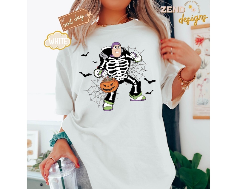 K&ouml;nnte beinhalten: Wei&szlig;es T-Shirt mit einem Halloween-Design. Die Grafik zeigt eine Figur in einem Skelettkost&uuml;m, die einen K&uuml;rbis h&auml;lt. Schwarze Flederm&auml;use und ein Spinnennetz vervollst&auml;ndigen das gruselige Design. Auf dem Shirt steht das Wort "WHITE".