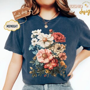 Puede incluir: Una camiseta azul marino con un dise&ntilde;o de ramo floral en tonos blancos, rosas y rojos. La camiseta tiene cuello redondo y mangas cortas. El texto "next day" y "Midnight" est&aacute; en la camiseta.