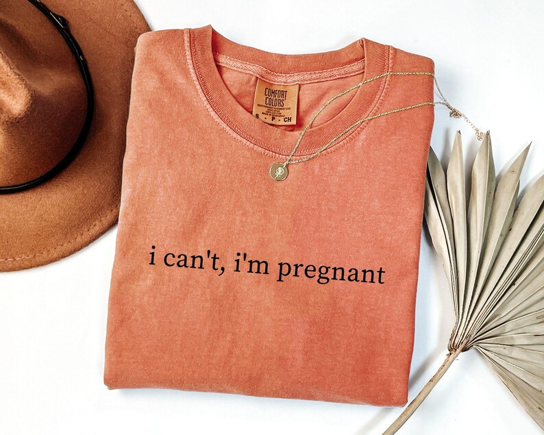 May include: Coral-colored t-shirt with the phrase "i can't, i'm pregnant" in black text. A gold necklace with a pendant and a brown hat are next to the shirt. A dried palm leaf is also in the image.