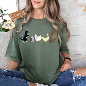 May include: Moss green t-shirt featuring a line of cartoon animals: a black and white cat, an owl, a chicken, a duck, and a bear. The shirt has a relaxed fit and is worn with blue jeans. The words "next day" and "MOSS" are on a tag.