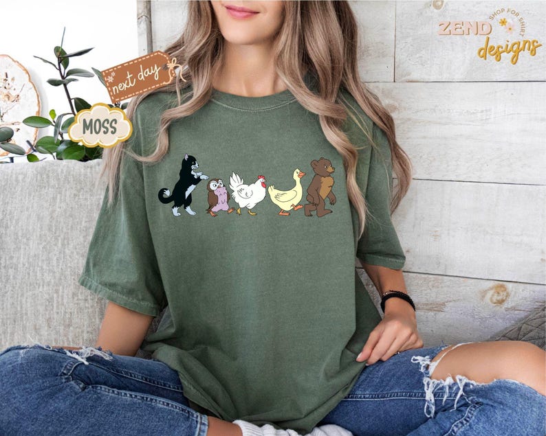 May include: Moss green t-shirt featuring a line of cartoon animals: a black and white cat, an owl, a chicken, a duck, and a bear. The shirt has a relaxed fit and is worn with blue jeans. The words "next day" and "MOSS" are on a tag.