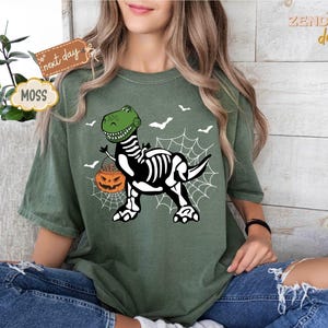 K&ouml;nnte beinhalten: Moosgr&uuml;nes T-Shirt mit einem Halloween-Motiv. Das Design zeigt einen gr&uuml;nen Dinosaurier mit Skelettk&ouml;rper, der einen K&uuml;rbis tr&auml;gt. Wei&szlig;e Flederm&auml;use und ein Spinnennetz erg&auml;nzen das Design. Das T-Shirt hat eine lockere Passform.