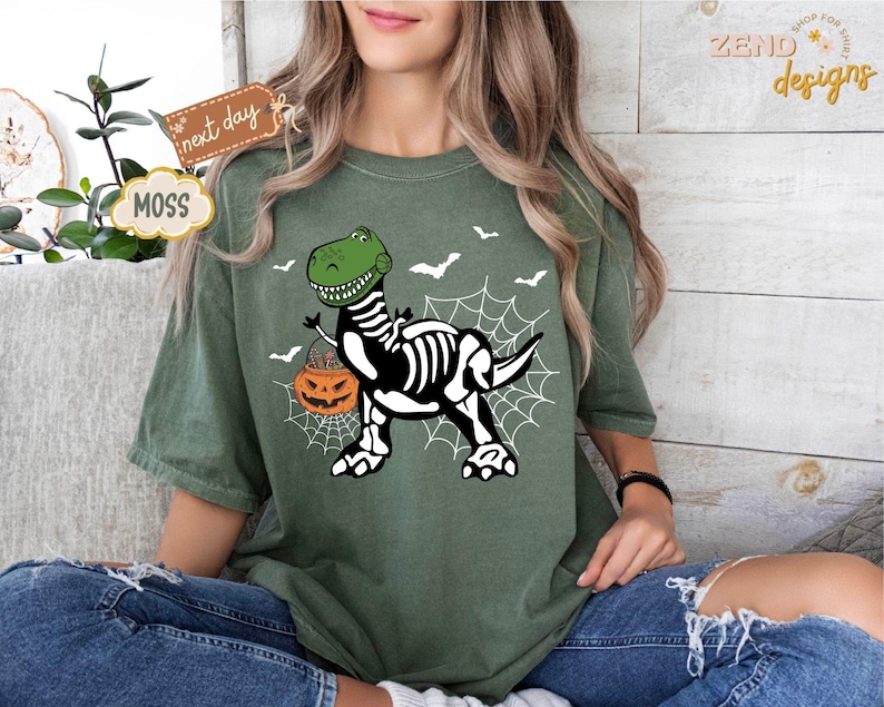K&ouml;nnte beinhalten: Moosgr&uuml;nes T-Shirt mit einem Halloween-Motiv. Das Design zeigt einen gr&uuml;nen Dinosaurier mit Skelettk&ouml;rper, der einen K&uuml;rbis tr&auml;gt. Wei&szlig;e Flederm&auml;use und ein Spinnennetz erg&auml;nzen das Design. Das T-Shirt hat eine lockere Passform.