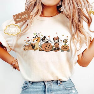 May include: Ivory-colored t-shirt featuring a fall-themed graphic with cartoon characters, including Mickey and Minnie Mouse, Goofy, and Pluto, posing with carved pumpkins. The shirt has the text "IVORY" and "next day" on it.