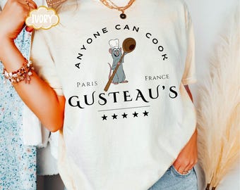 May include: A white t-shirt with a black graphic design featuring a cartoon rat chef holding a wooden spoon. The graphic says "Anyone Can Cook" in a circle around the rat, with "Paris" and "France" on either side of the rat. Below the rat, it says "Gusteau's" with five stars.