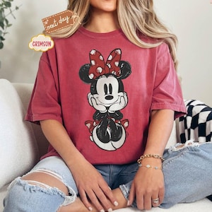 May include: A red t-shirt with a vintage-style graphic of Minnie Mouse wearing a red bow with white polka dots. The shirt has a relaxed fit and short sleeves.