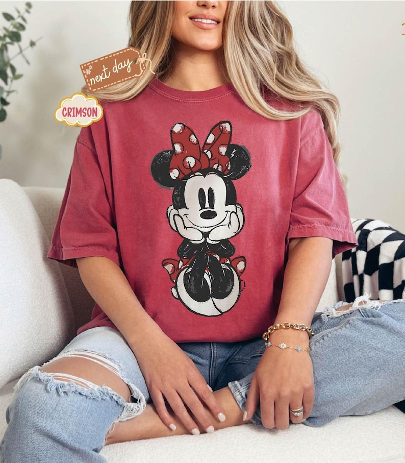 May include: A red t-shirt with a vintage-style graphic of Minnie Mouse wearing a red bow with white polka dots. The shirt has a relaxed fit and short sleeves.