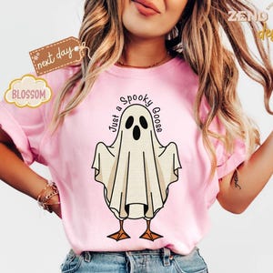 May include: A light pink t-shirt featuring a cartoon ghost with the text "Just a Spooky Goose". The ghost has black eyes and a beak, with orange feet. The shirt is a casual style.