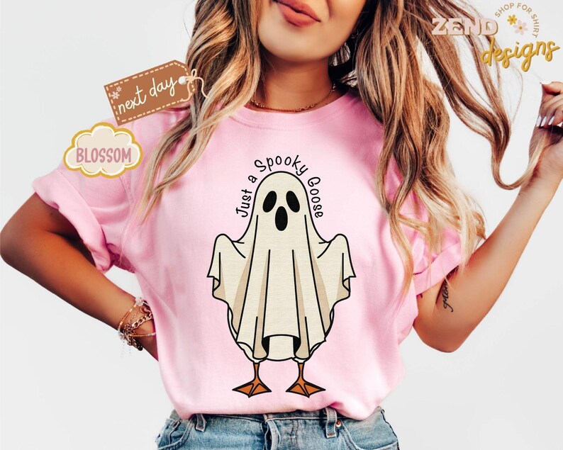 May include: A light pink t-shirt featuring a cartoon ghost with the text "Just a Spooky Goose". The ghost has black eyes and a beak, with orange feet. The shirt is a casual style.