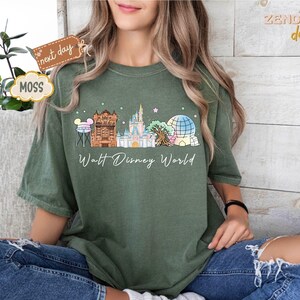 May include: A moss green t-shirt featuring a colorful illustration of Walt Disney World landmarks, including Cinderella's castle and Spaceship Earth. The words "Walt Disney World" are written in a cursive font below the graphic. The shirt is worn by a person.