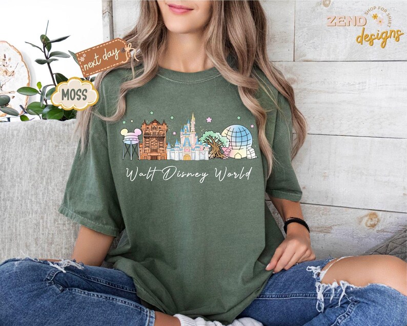 May include: A moss green t-shirt featuring a colorful illustration of Walt Disney World landmarks, including Cinderella's castle and Spaceship Earth. The words "Walt Disney World" are written in a cursive font below the graphic. The shirt is worn by a person.