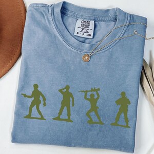 Puede incluir: Una camiseta azul Comfort Colors con cuatro siluetas de soldados de juguete verdes. Un collar dorado con un colgante est&aacute; drapeado sobre la camisa. Un sombrero marr&oacute;n y tallos de plantas secas est&aacute;n en el fondo.
