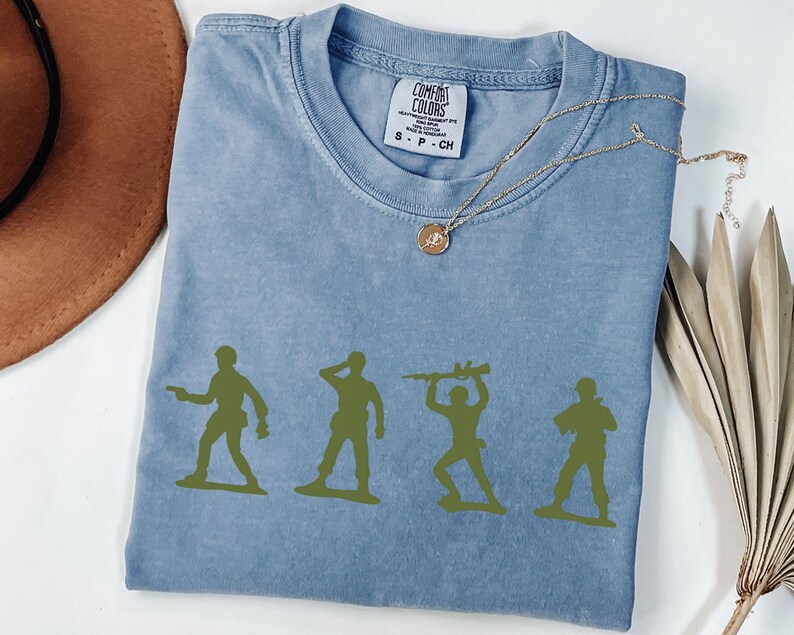 Puede incluir: Una camiseta azul Comfort Colors con cuatro siluetas de soldados de juguete verdes. Un collar dorado con un colgante est&aacute; drapeado sobre la camisa. Un sombrero marr&oacute;n y tallos de plantas secas est&aacute;n en el fondo.