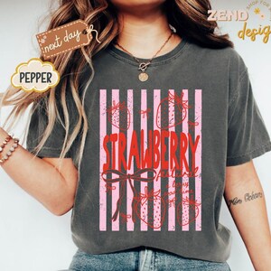 May include: Dark gray t-shirt featuring a pink and white striped design with strawberries and the text "STRAWBERRY Festival". The shirt has a relaxed fit and short sleeves. The word "PEPPER" is on a cloud-shaped tag.